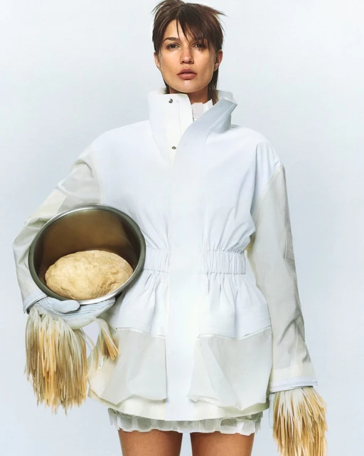 Woman in white jacket holds bowl of dough in studio. Minimalist fashion shot with neutral tones and unusual food styling concept.