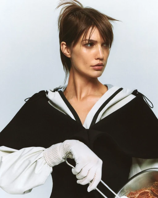 Portrait of a fair-skinned adult woman posing in a black and white top holding a metal bowl in white gloves against a white background.