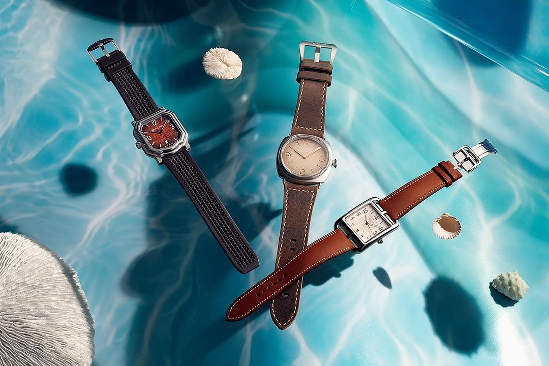 Three wristwatches with varying brown leather straps are showcased against a textured, light-blue backdrop with sea-themed accents.