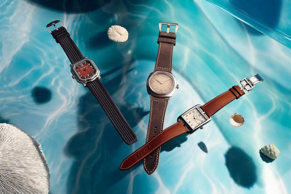 Three wristwatches with varying brown leather straps are showcased against a textured, light-blue backdrop with sea-themed accents.