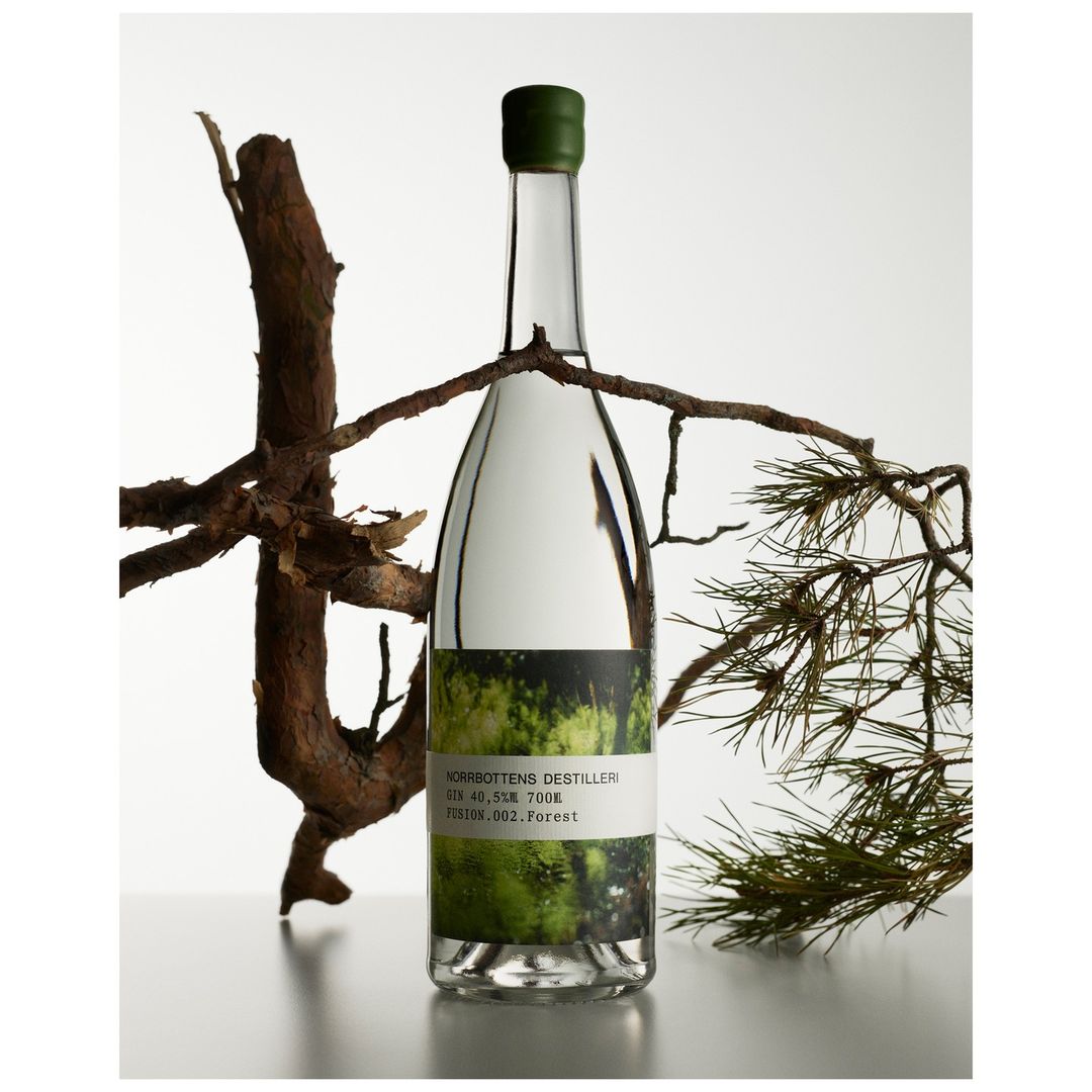 Bottle of clear liquor with green accents displayed with tree branches against white background. The label features forest imagery.