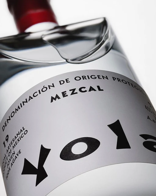 Close-up of a clear glass mezcal bottle with a white label and black text, featuring the brand name 'Volans' prominently.