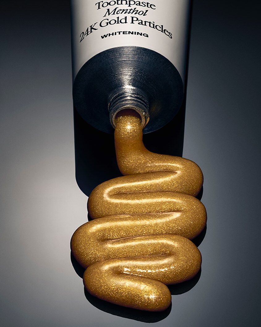 Close-up shot of a tube of toothpaste with gold toothpaste being squeezed out in a wavy pattern on a dark gray background.