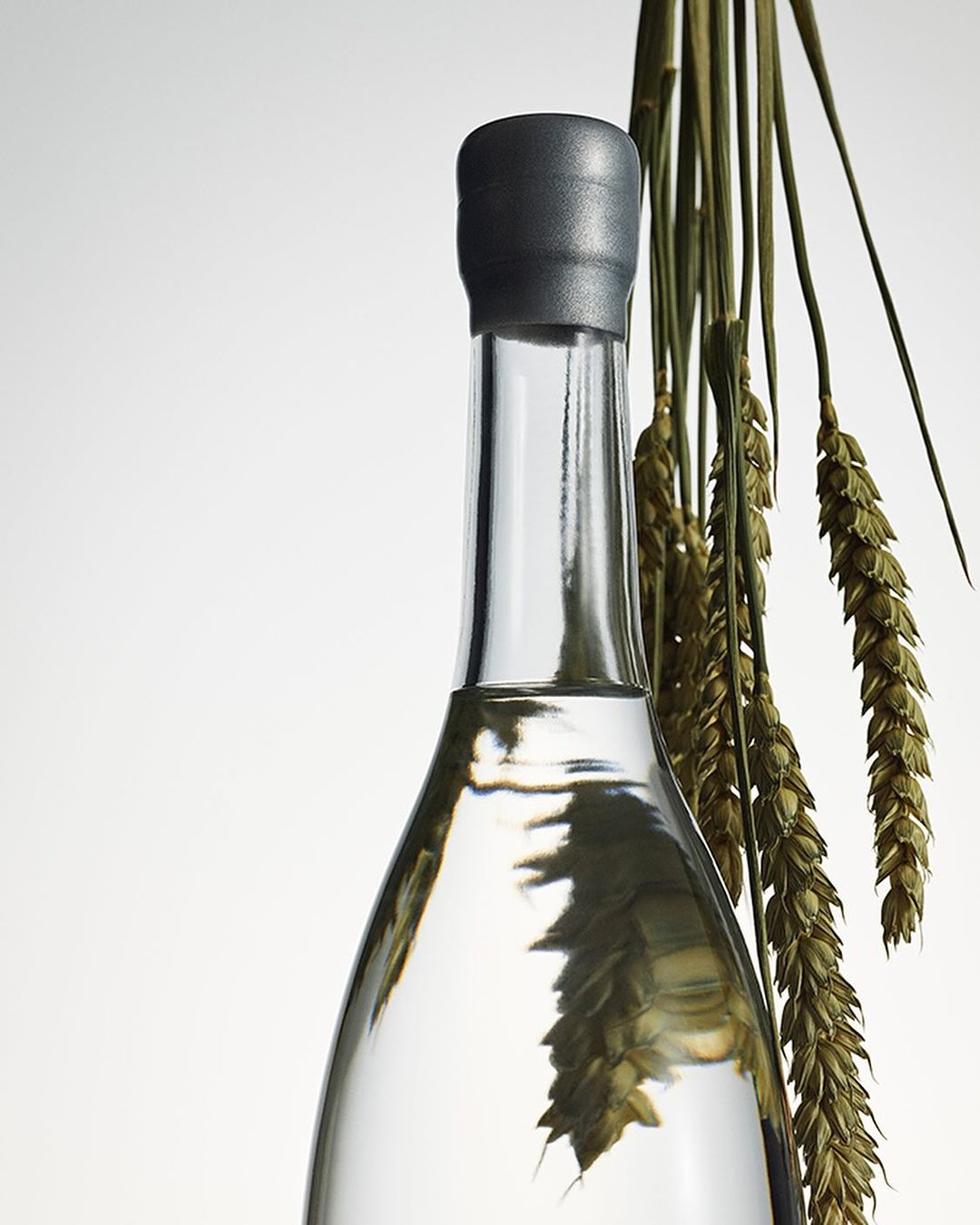 Close-up of a clear glass bottle with a gray cap, filled with a translucent liquid, and decorated with dried wheat stalks.