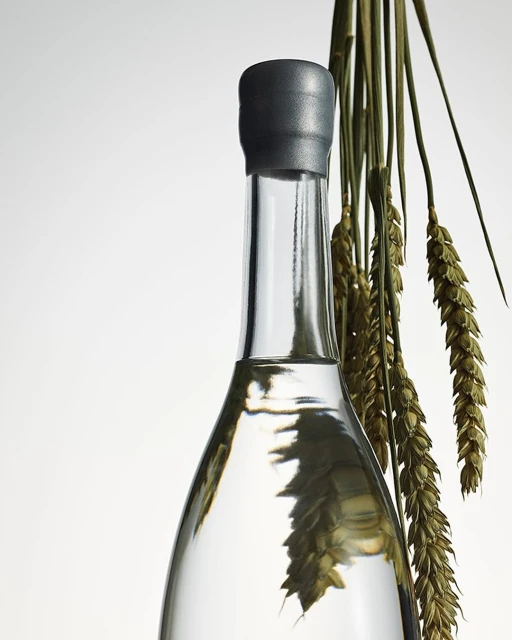 Close-up of a clear glass bottle with a gray cap, filled with a translucent liquid, and decorated with dried wheat stalks.