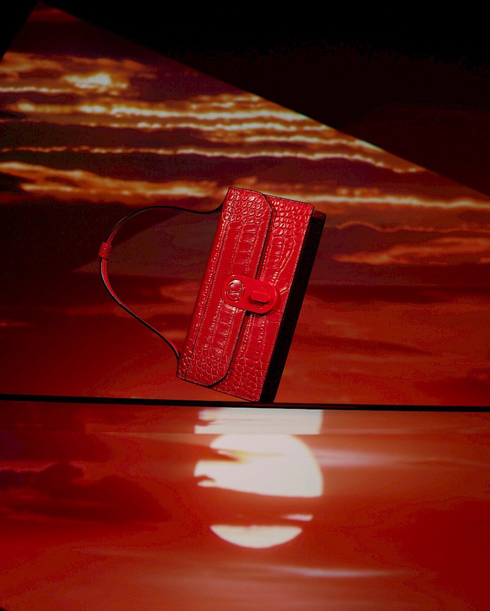 Fashion still life: a vibrant red, crocodile-textured clutch purse positioned on a reflective surface with a dreamy, cloud-filled backdrop.