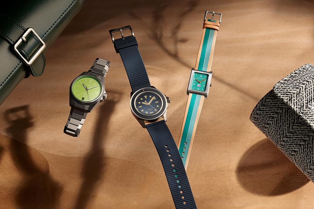 Three Unimatic wristwatches are displayed on a tan surface, each featuring unique band styles: metal, rubber, and striped fabric.