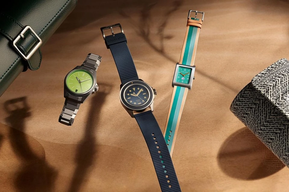Three Unimatic wristwatches are displayed on a tan surface, each featuring unique band styles: metal, rubber, and striped fabric.