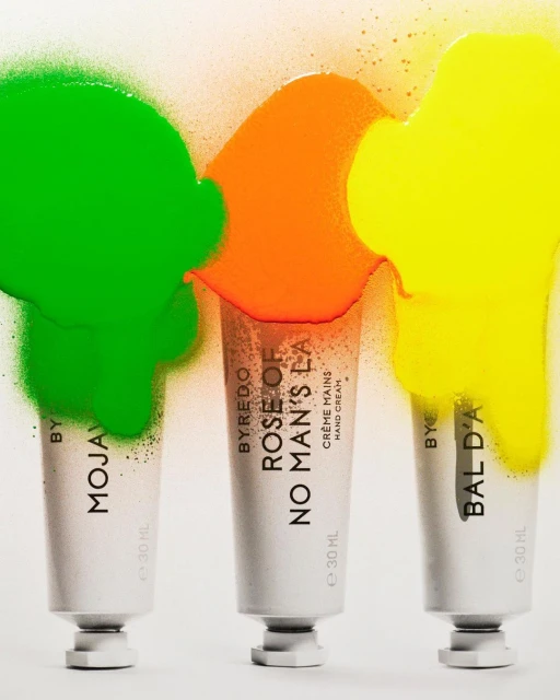 Three tubes of Byredo hand cream with green, orange and yellow paint-like blobs against a white backdrop in a product shot.