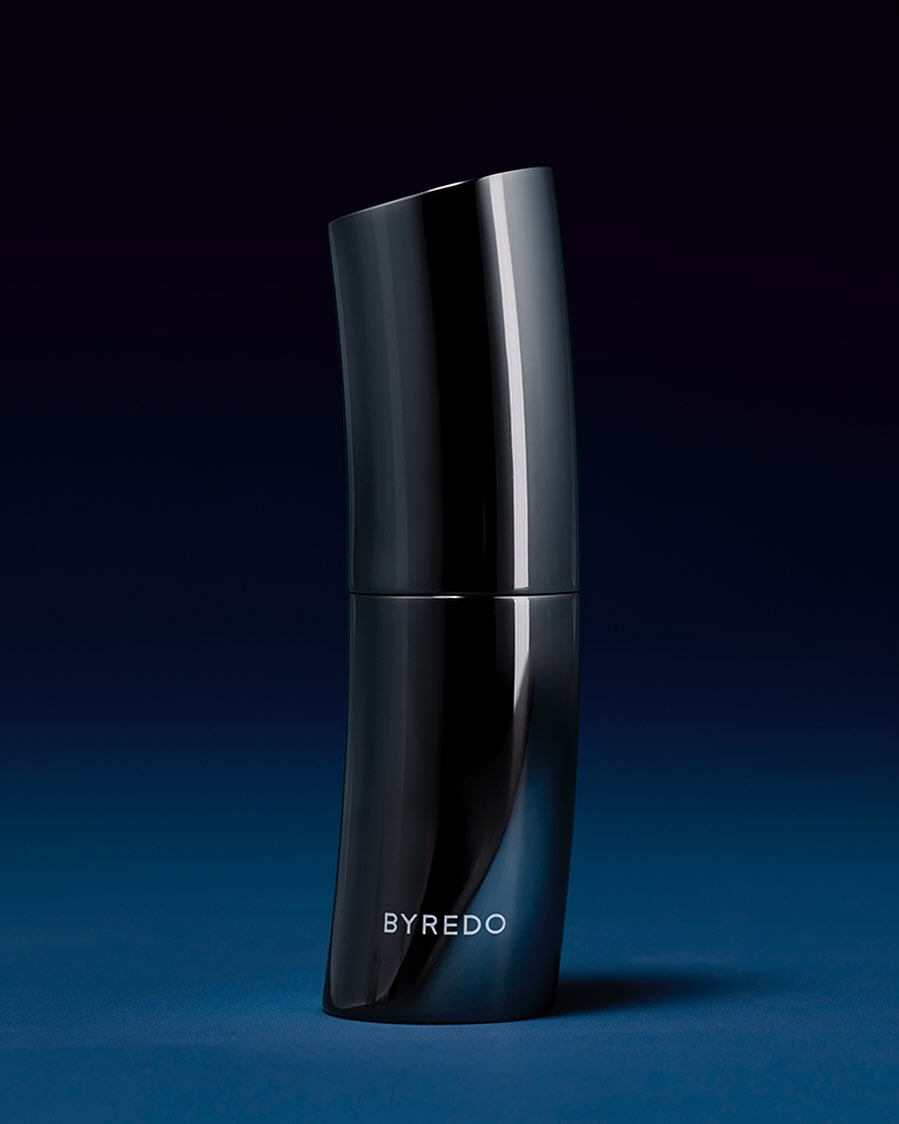 Sleek, curved dark gray perfume bottle by Byredo on a dark blue surface with subtle reflections, minimal modern product shot, beauty.