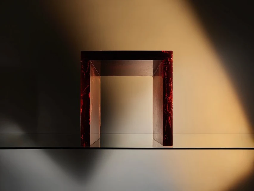 A modern display featuring a unique U-shaped object with a red marble-like finish, showcased on a reflective glass surface against a gradient backdrop.