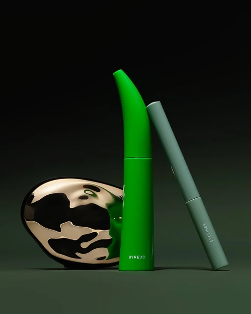 Still life shot of green Byredo mascara, grey eyeliner, and gold cosmetic compact against a dark green backdrop in a studio setting.