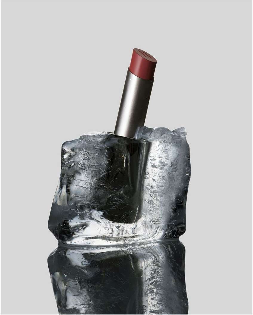 Product shot of a red lipstick in a silver tube, embedded in a block of ice, set against a plain gray background with a reflective surface.
