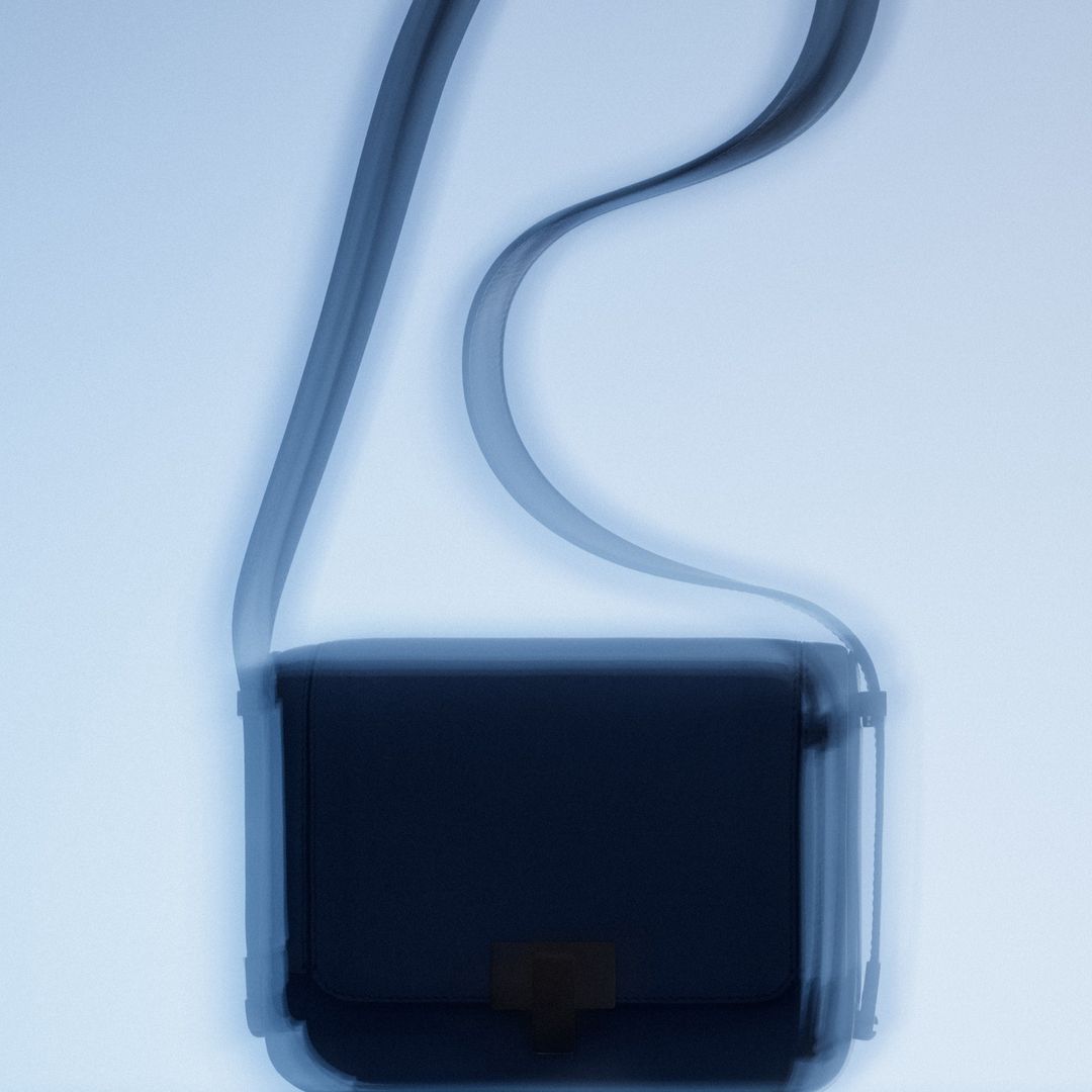 Monochromatic x-ray of a dark cross-body bag with a thin strap, laid flat on a light blue surface, showing inner details.