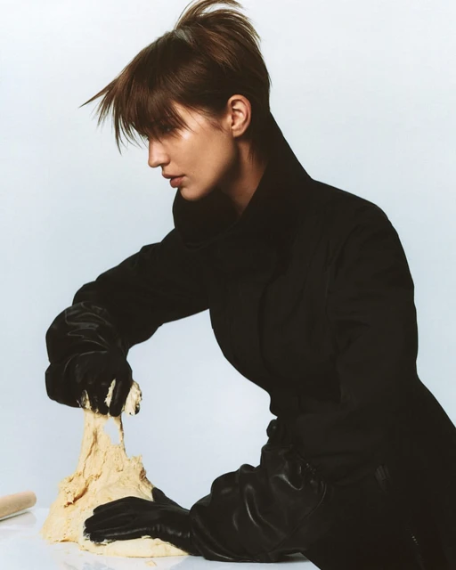 Woman in a black coat kneading dough, wearing gloves, against a plain background in a studio environment, culinary or beauty editorial shot.