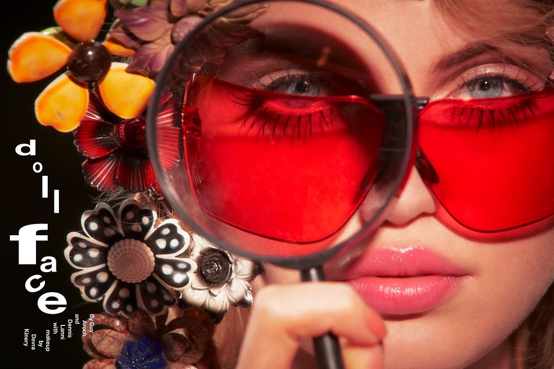 Close-up of a woman with red sunglasses, floral headpiece, and fair skin, holding a magnifying glass, showcasing fashion and beauty elements.
