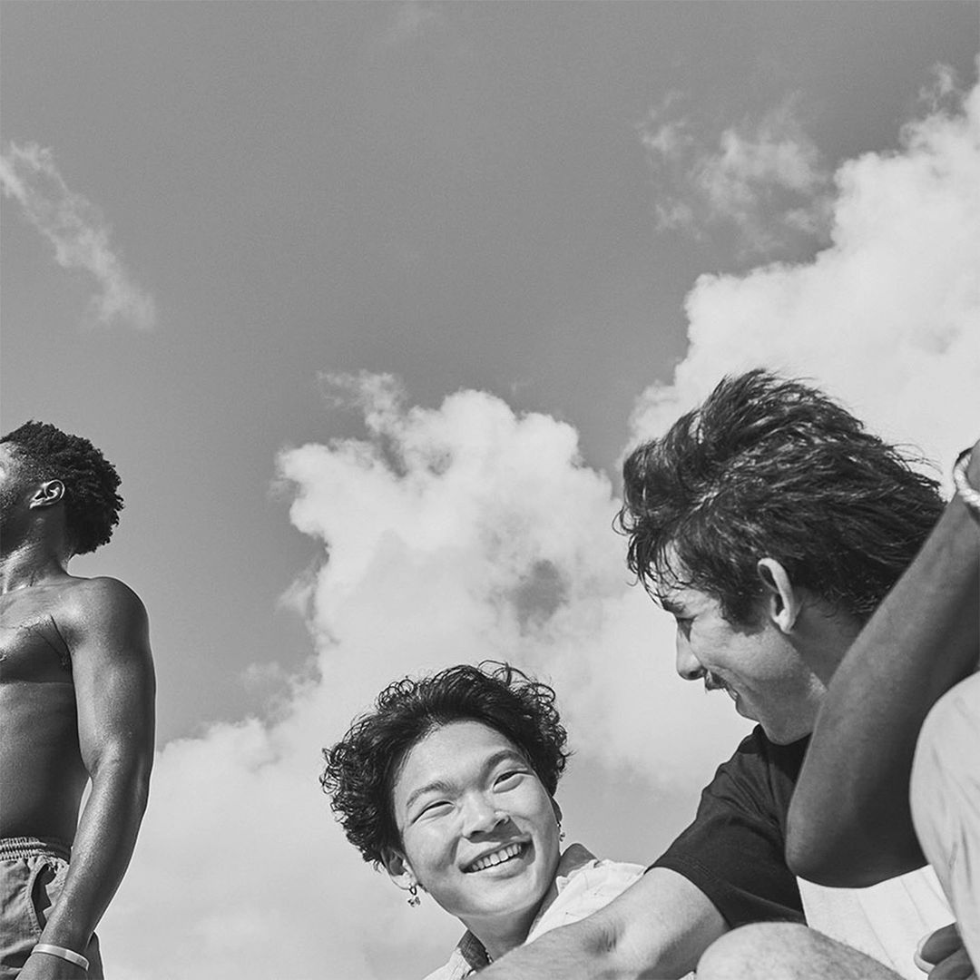 Low-angle, black and white portrait of a diverse group of young adults smiling and looking upwards against a cloudy sky backdrop.