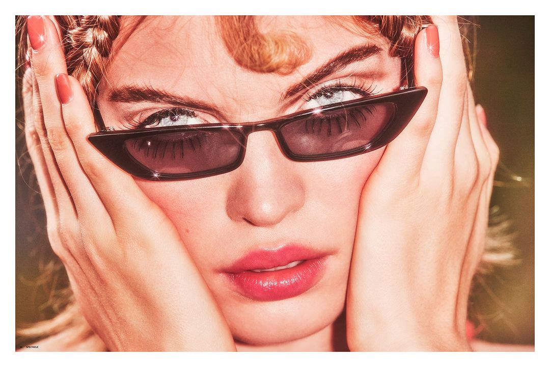 Close-up of a light-skinned woman wearing sunglasses, with her hands near her face, and her hair in braids and loose strands.