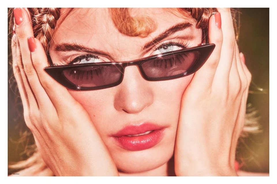 Close-up of a light-skinned woman wearing sunglasses, with her hands near her face, and her hair in braids and loose strands.