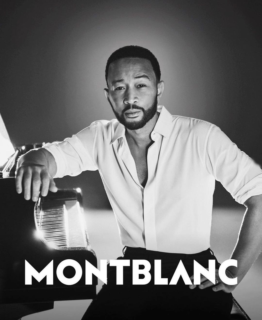 Black and white portrait of John Legend with a piano, wearing a white button-down shirt and black pants, with the MONTBLANC logo.