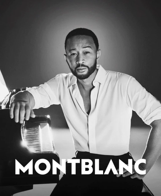 Black and white portrait of John Legend with a piano, wearing a white button-down shirt and black pants, with the MONTBLANC logo.