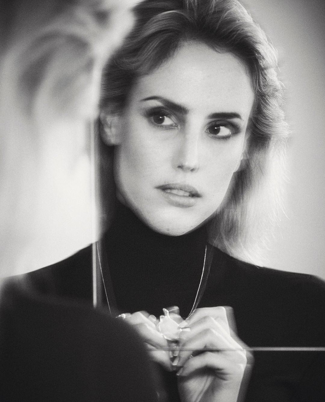 Black and white portrait of a woman with blonde hair looking in a mirror, wearing a black turtleneck sweater and a necklace.
