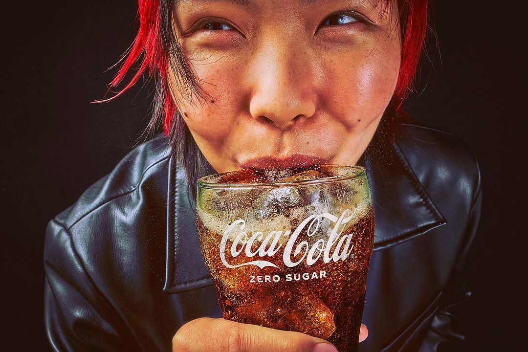 Close up of an Asian adult female drinking a Coca-Cola Zero Sugar with ice from a glass, wearing a black leather jacket.