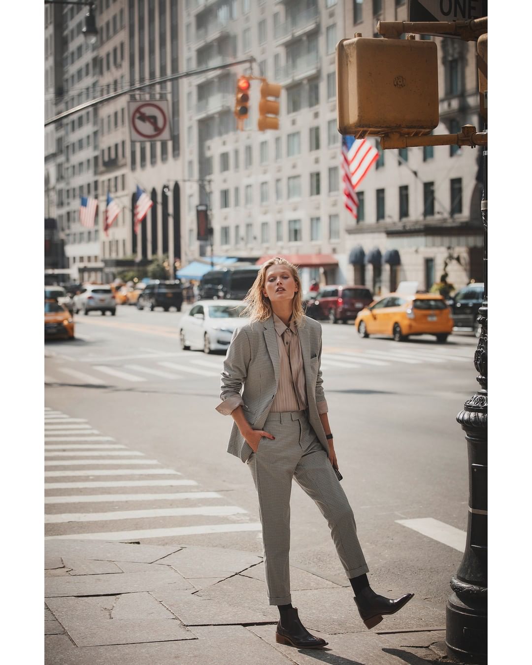 Fashion photography of a blonde woman in a grey suit standing in an urban setting on a city street with buildings and traffic.