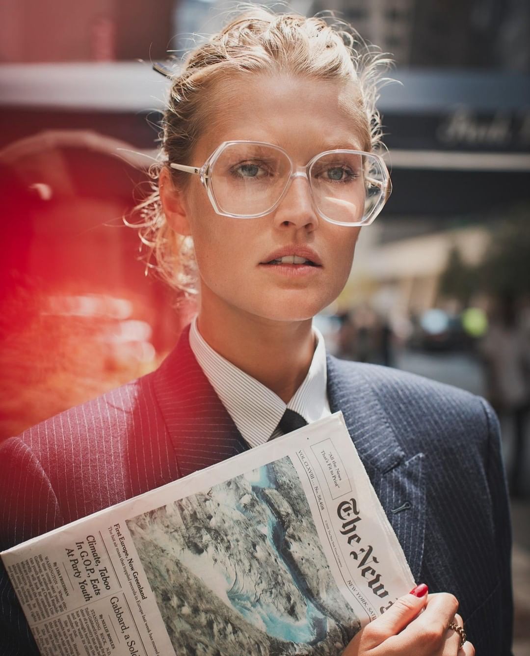 Adult woman poses in front of city buildings while holding a newspaper, wearing clear-framed glasses and a striped shirt and suit.