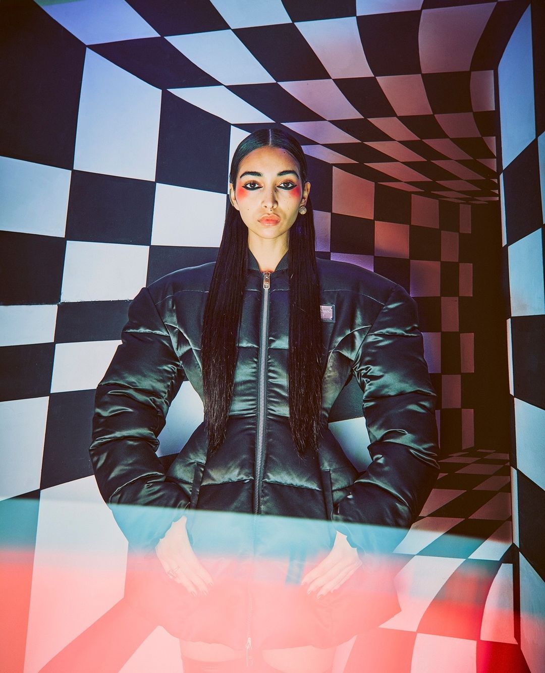 Woman in an oversized puffer jacket poses in a studio with a black-and-white checkerboard pattern and red and blue lighting accents.