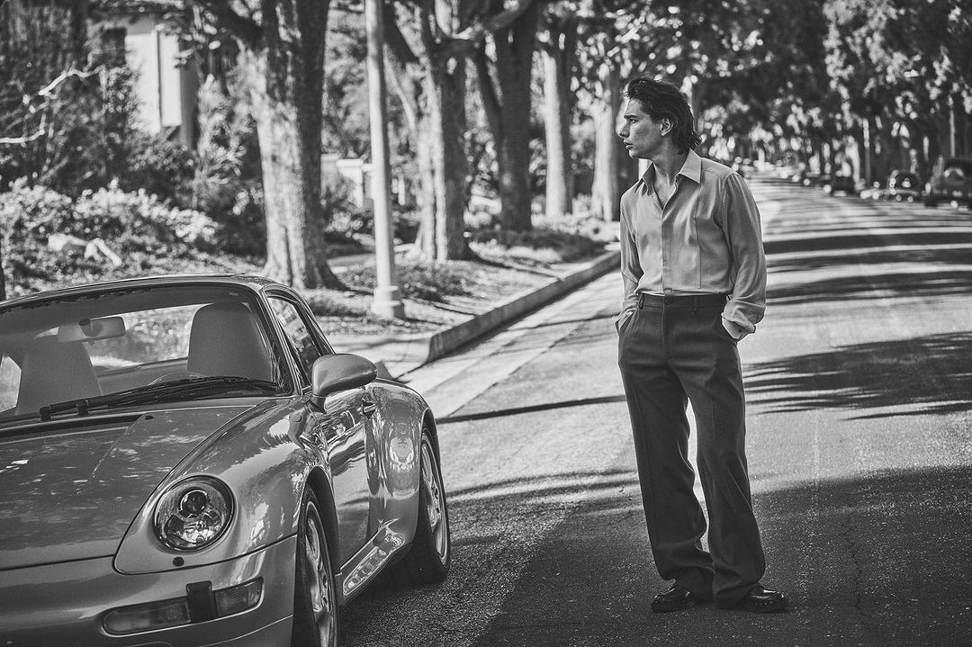 Man standing on a residential street next to a parked vintage sports car, captured by Guy Aroch in New York.