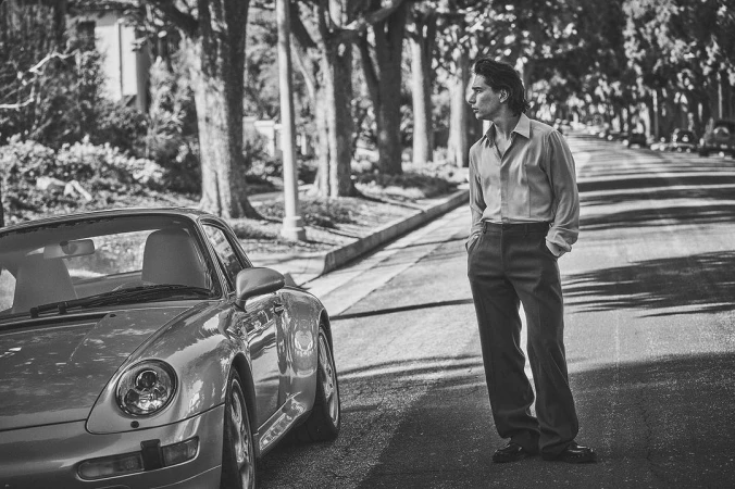 Man standing on a residential street next to a parked vintage sports car, captured by Guy Aroch in New York.