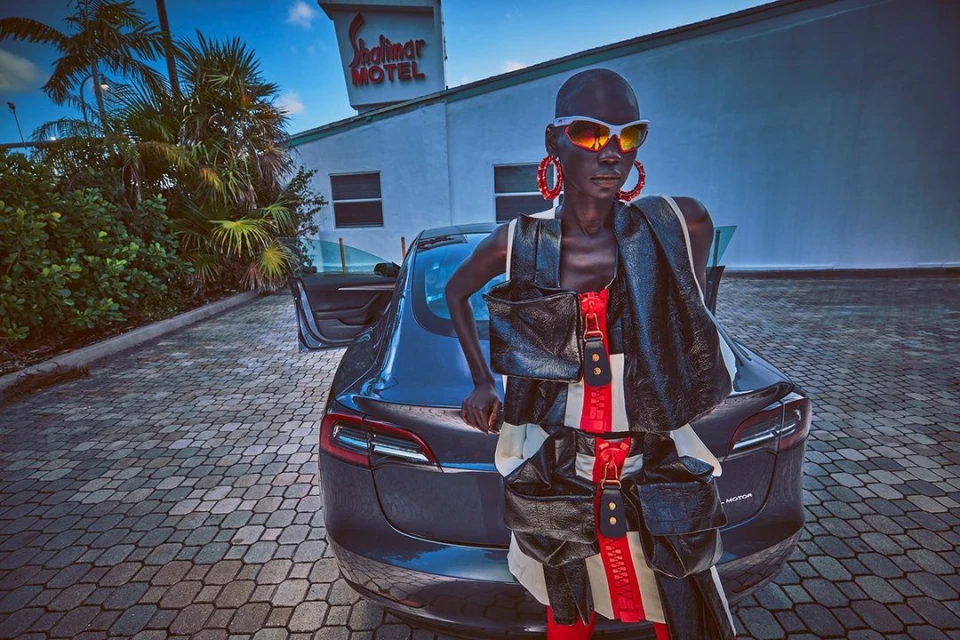 Woman in avant-garde fashion poses with a dark grey electric car in front of a vintage motel with palm trees in the background.