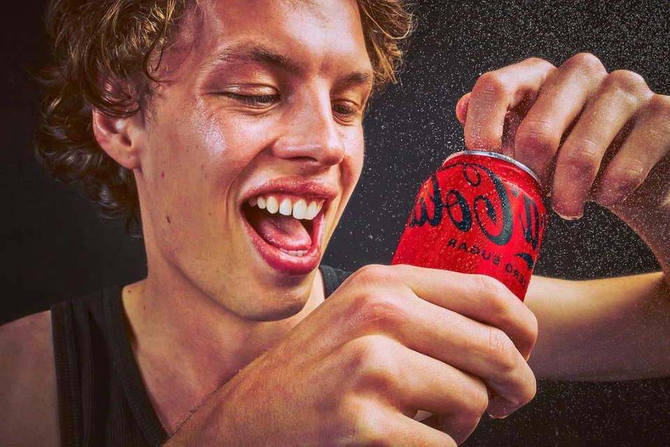Excited young man opens a bright red can of Coca-Cola Zero Sugar with water droplets in a close-up shot on a dark background.