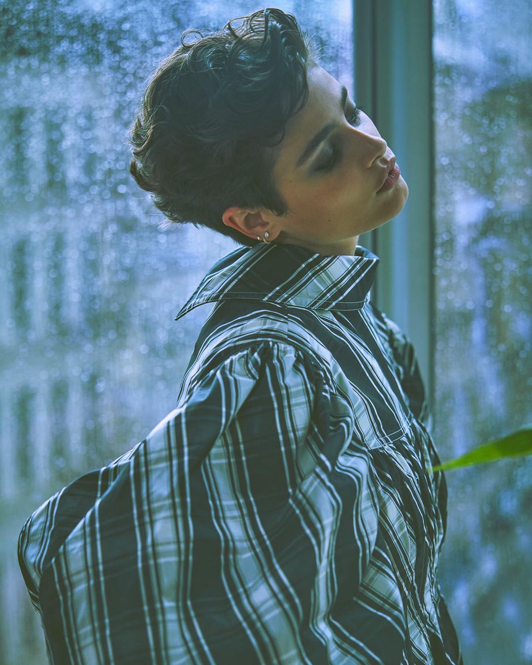 Portrait of a person with short dark hair wearing a plaid blouse against a rainy window backdrop.
