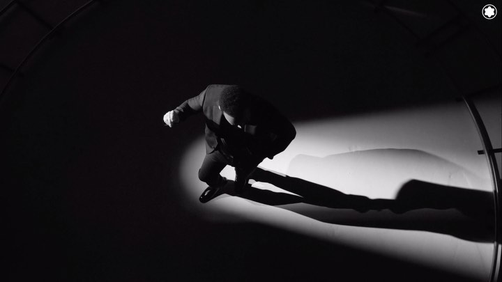Monochrome, low-angle action shot of an adult man leaping in a dark suit against a stark background with dramatic lighting and visible shadow.