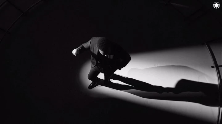 Monochrome, low-angle action shot of an adult man leaping in a dark suit against a stark background with dramatic lighting and visible shadow.