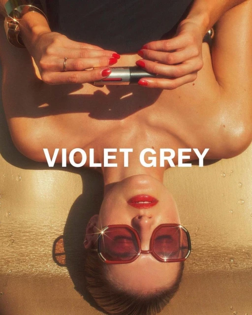 Adult woman with red lipstick and sunglasses is lying down holding a cosmetic product on a tan surface, fashion and beauty concept.