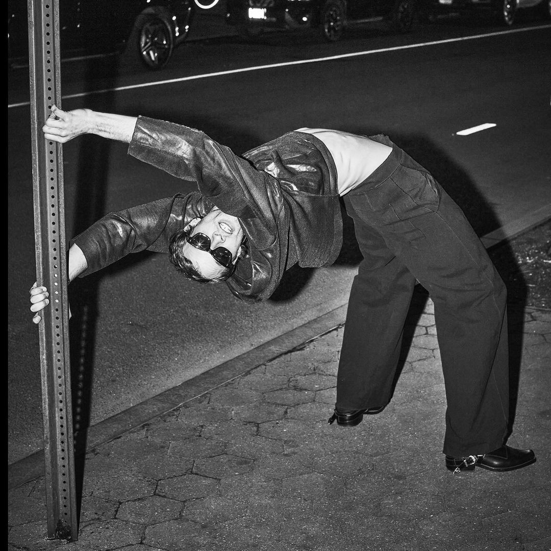 An adult wearing a leather jacket leans backwards, supported by a metal post on a city street at night, vehicles in background.