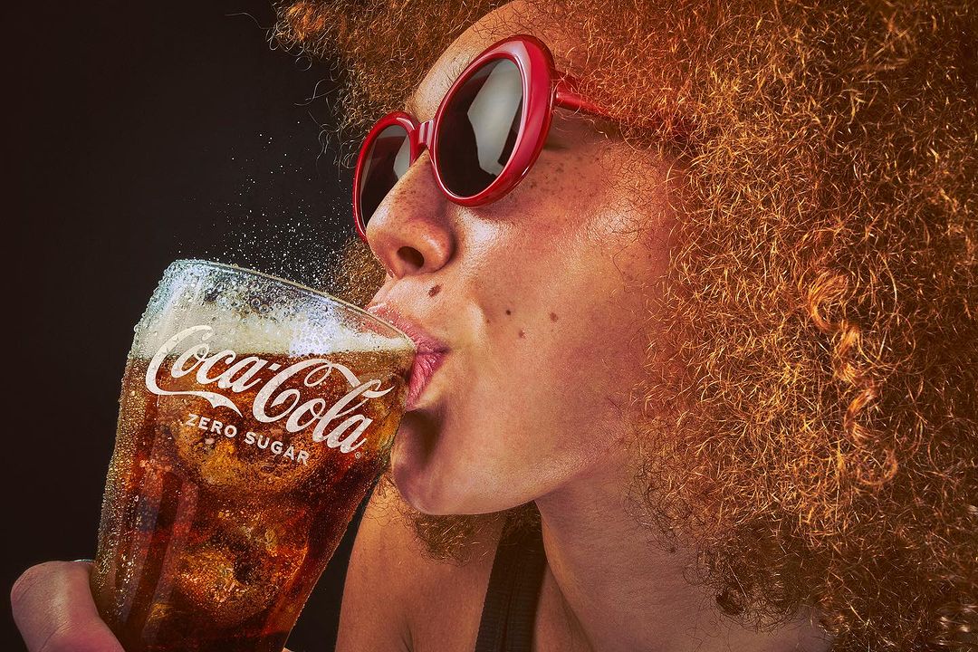 Woman drinking Coca-Cola Zero Sugar in a close-up studio shot. She wears red sunglasses and has curly auburn hair against a dark backdrop.