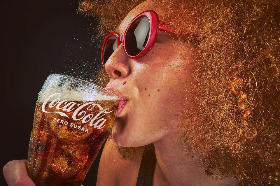 Woman drinking Coca-Cola Zero Sugar in a close-up studio shot. She wears red sunglasses and has curly auburn hair against a dark backdrop.