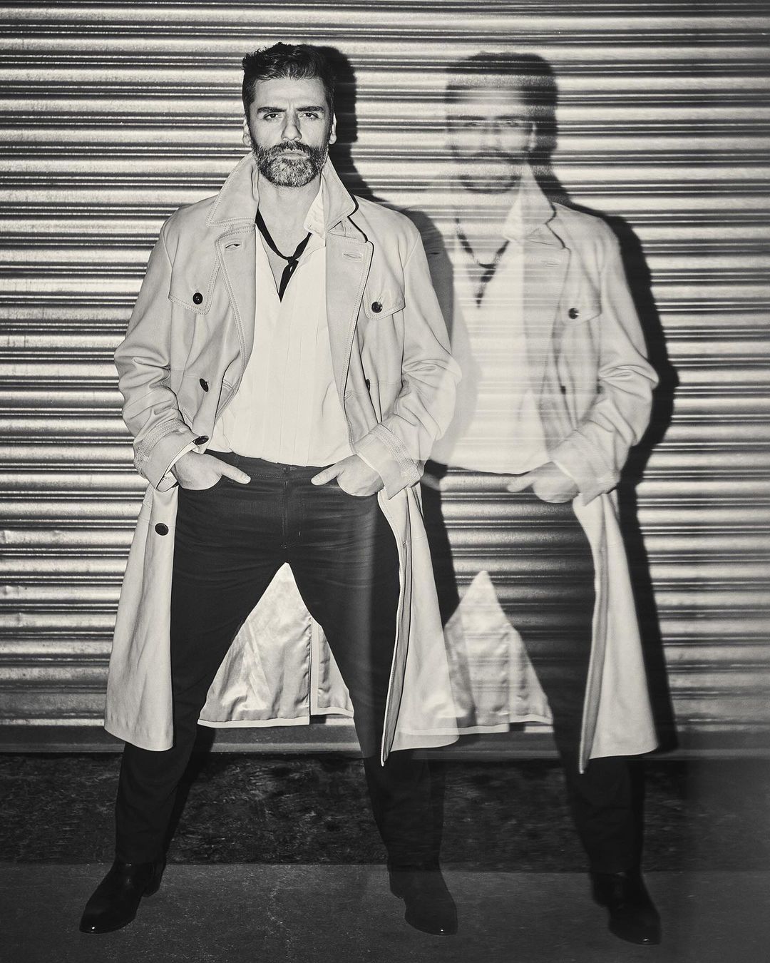 Monochrome portrait of a stylish man in a trench coat standing against a corrugated metal wall with a blurred, ghost-like afterimage.