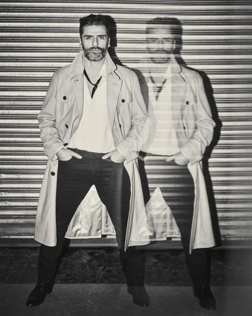 Monochrome portrait of a stylish man in a trench coat standing against a corrugated metal wall with a blurred, ghost-like afterimage.
