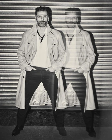 Monochrome portrait of a stylish man in a trench coat standing against a corrugated metal wall with a blurred, ghost-like afterimage.
