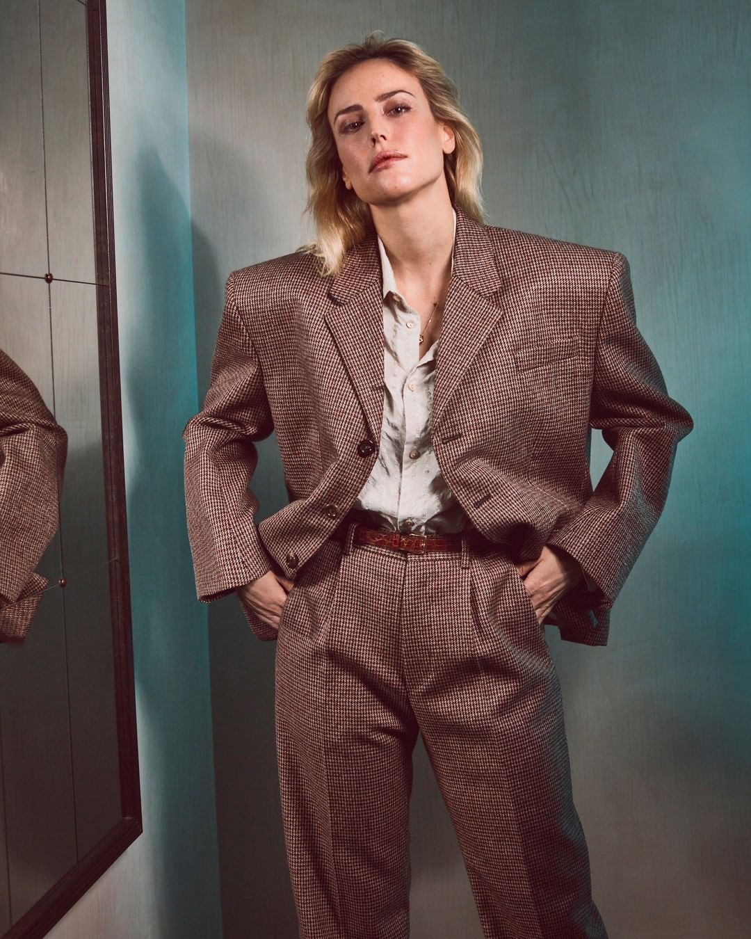 Blonde woman poses indoors wearing a brown suit and light button-down shirt. She has her hands in her pockets.