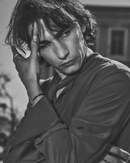 Monochrome portrait of a young adult man with dark hair, wearing a shirt, with his hand near his face in an outdoor setting.