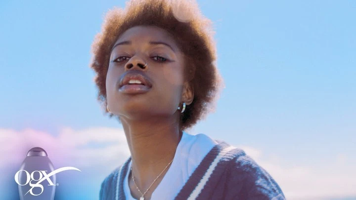 Young Black woman gazes upward with Afro hair, wearing a cardigan against a blue sky, promoting OGX hair product.