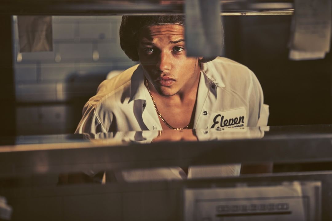 Eye-level shot of a young man with a neutral expression, wearing a uniform shirt with 'Eleven' embroidered on it, indoors.