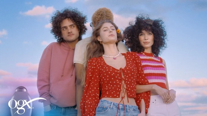 Four models pose outdoors against a blue, cloudy sky promoting OGX hair care products. Red and pink tops contrast with denim and white pants.