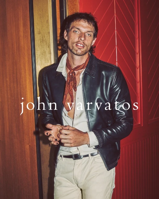 Man in leather jacket, neck scarf, and light pants poses against a paneled wall with a door. Fashion advertisement for John Varvatos.